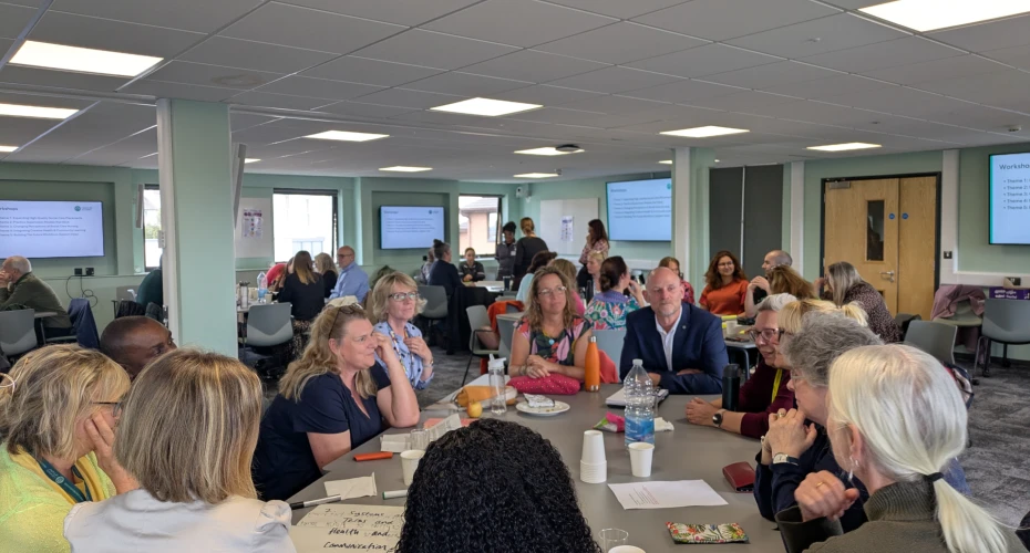 Photo taken of a large room, with roughly 5 desks with a number of people sat around each desk. There are 4 tv screens on the wall in the background and you can see three windows in the distance. On the desks there are various pieces of paper, pens and water bottles.