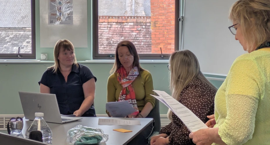 Image of 4 people near a desk in a room. Three people are sat around the table with one person stood reading a piece of paper.
