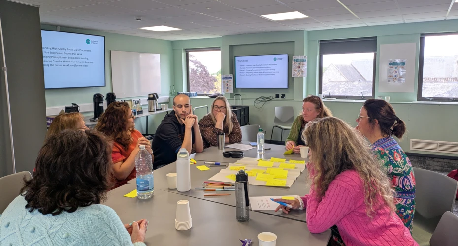 Group of 8 people sat around desks in a room, in discussion with each other. There are numerous post it notes stuck to a large peice of paper, various bottles on the table, two screens on the wall in the background and three windows.